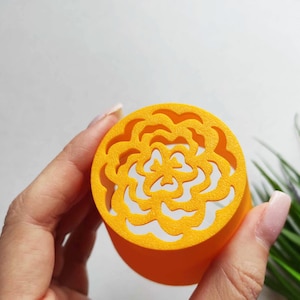May include: A circular, orange container featuring a laser-cut floral design on its lid. The lid's detailed pattern exposes a white interior. The container is held against a soft, white backdrop, with green and purple plants in the background.