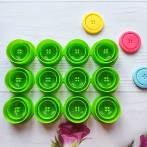 Può includere: Un set di 12 stampi per bottoni in plastica verde, disposti in una griglia 4x3. Ci sono tre bottoni aggiuntivi, uno giallo, uno rosa e uno blu.