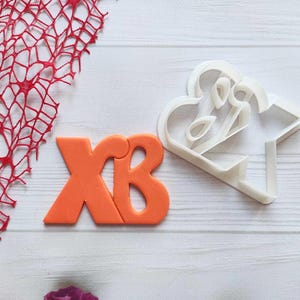 May include: Two cookie cutters, one shaped like the Cyrillic letters "XB" in orange and the other shaped like a stylized floral design in white.