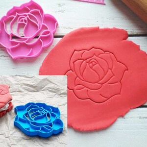 May include: Three rose-shaped cookie cutters, one pink, one blue, and one red. The red cutter is used to cut out a rose shape from a red piece of modeling clay.