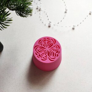 Könnte beinhalten: Ein kreisförmiges, leuchtend rosa Objekt mit einem vertieften, komplizierten blumenartigen Muster auf der Oberseite. Der Gegenstand hat eine glatte, massive zylindrische Basis und eine matte Oberfläche mit einem symmetrischen Design.