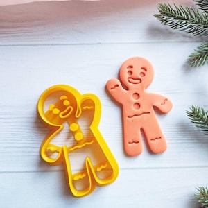 Puede incluir: Dos cortadores de galletas de hombre de jengibre, uno amarillo y otro naranja. El cortador amarillo es un contorno detallado de un hombre de jengibre con una cara sonriente, los brazos extendidos y un botón en el pecho. El cortador naranja es un hombre de jengibre sólido con una cara sonriente, los brazos extendidos y un botón en el pecho.