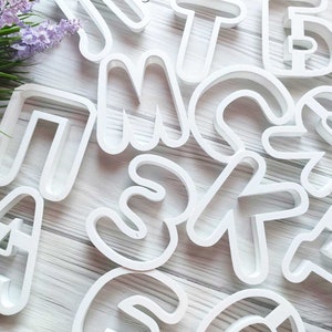 May include: White plastic cookie cutters shaped like letters of the alphabet. The letters are arranged in a random pattern on a white wooden surface.