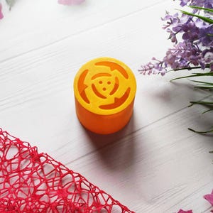 May include: A bright orange round silicone mold with a flower design. The mold is on a white surface with a red mesh fabric and purple flowers.