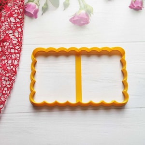 Puede incluir: Un cortador de galletas rectangular amarillo con un borde festoneado y un divisor vertical. El cortador está sobre una superficie de madera blanca, con una tela de malla roja y rosas rosas en el fondo. Esta herramienta es para hornear.