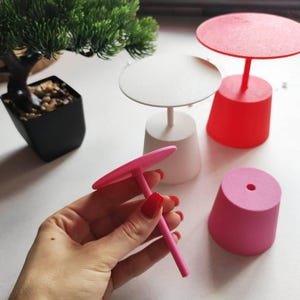 Peut inclure: Un ensemble de petits présentoirs circulaires avec des bases coniques en blanc, rouge et rose. Un présentoir rose est tenu à la main, montrant un plateau plat fixé à une fine tige verticale qui s'insère dans la base. Les supports sont mats.