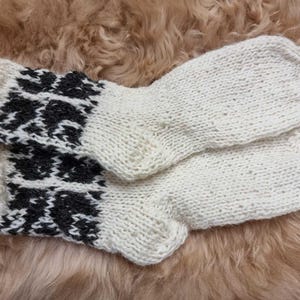 K&ouml;nnte beinhalten: Ein Paar wei&szlig;e Stricksocken mit einem schwarz-wei&szlig;en geometrischen Muster am B&uuml;ndchen. Die Socken sind aus Wolle und weich und warm.