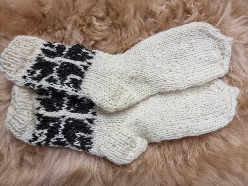 K&ouml;nnte beinhalten: Ein Paar wei&szlig;e Stricksocken mit einem schwarz-wei&szlig;en geometrischen Muster am B&uuml;ndchen. Die Socken sind aus Wolle und weich und warm.