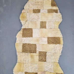 Pode incluir: Um tapete de pele de carneiro patchwork em um esquema de cores bege e branco. O tapete é feito de diferentes pedaços de pele de carneiro costurados juntos para criar um padrão xadrez. O tapete tem uma aparência e sensação naturais e rústicas.