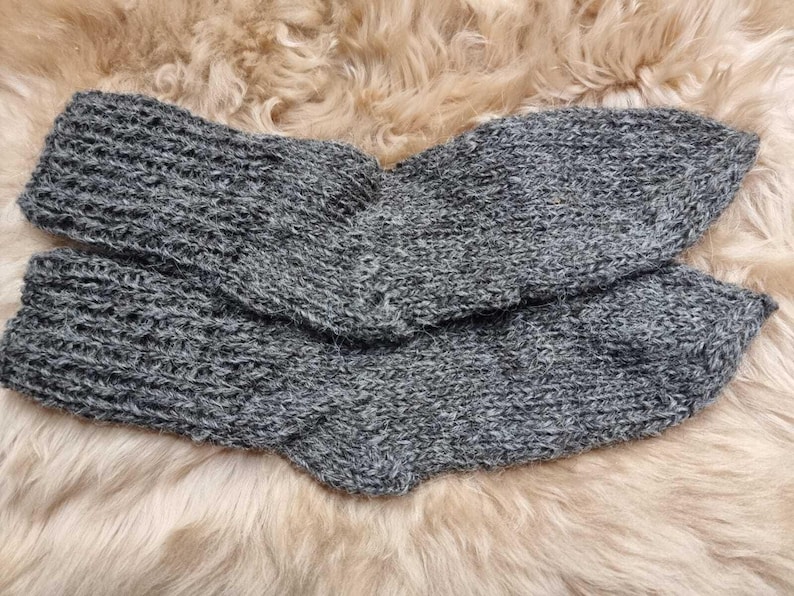 K&ouml;nnte beinhalten: Ein Paar graue Stricksocken mit einer flauschigen Textur. Die Socken sind in der H&auml;lfte gefaltet und auf einer flauschigen, hellbraunen Oberfl&auml;che ausgelegt.