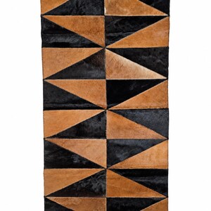 Puede incluir: Una alfombra rectangular construida con paneles de piel de vaca cosidos en patrones triangulares alternos de color negro y bronceado. La superficie presenta un diseño geométrico repetitivo con costuras visibles que conectan cada pieza.