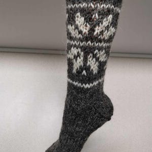K&ouml;nnte beinhalten: Graue Wollsocken mit einem wei&szlig;en Schneeflockenmuster. Die Socken sind gestrickt und haben eine flauschige Textur.