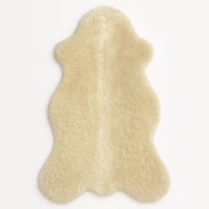 Könnte beinhalten: Ein cremefarbener Schaffellteppich mit einer einzigartigen, organischen Form. Der Teppich hat eine weiche, flauschige Textur und besteht aus natürlicher Wolle. Der Teppich hat eine neutrale Farbe und eignet sich daher für verschiedene Einrichtungsstile.