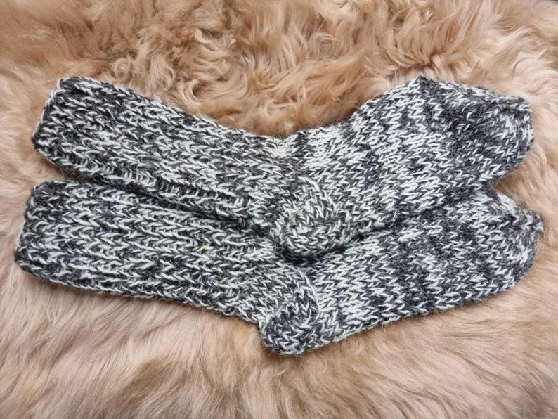 K&ouml;nnte beinhalten: Ein Paar kuschelige, gestrickte Socken in einem grau-wei&szlig;en Farbschema. Die Socken sind aus einem weichen, flauschigen Garn gefertigt und haben ein strukturiertes Strickmuster.