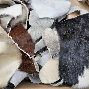 Puede incluir: Un montón de retazos de cuero de varios colores y texturas. Las piezas incluyen tonos de marrón, blanco, negro y gris, con pelo visible y superficies lisas. El cuero parece ser de diferentes pieles de animales.