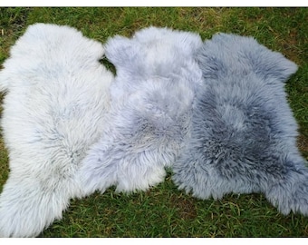 Puede incluir: Tres alfombras de piel de oveja en diferentes tonos de gris. Las alfombras están dispuestas sobre una superficie de hierba verde.