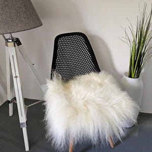 May include: A white faux fur throw rug placed on a black mesh chair. The chair is in front of a white wall and a gray lamp with a white tripod stand.