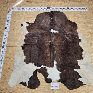 Könnte beinhalten: Ein brauner und weißer Kuhfellteppich mit einer natürlichen, haarigen Textur. Der Teppich misst etwa 100 cm in der Länge und 150 cm in der Breite. Der Teppich ist mit der Nummer 35-005 gekennzeichnet.