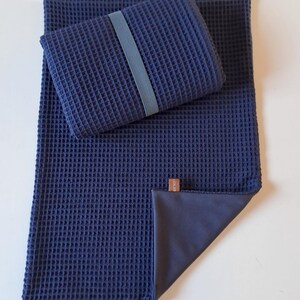Può includere: Una coperta in tessuto a nido d'ape blu scuro con un cuscino in tessuto a nido d'ape blu scuro. Il cuscino ha un elastico blu scuro intorno. La coperta ha un retro in tessuto blu scuro.