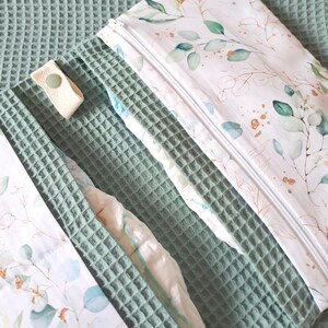 Peut inclure: Un sac à couches blanc et vert à imprimé floral avec une fermeture éclair, ouvert pour révéler une couche. Le sac est posé sur une surface texturée vert clair. Une sangle assortie avec une fermeture à bouton est visible.
