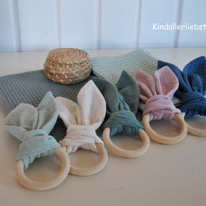 Op de afbeelding: Vijf houten bijtringen met stoffen konijnenoortjes in verschillende kleuren, waaronder lichtblauw, wit, teal, roze en blauw. De ringen liggen op een wit oppervlak.