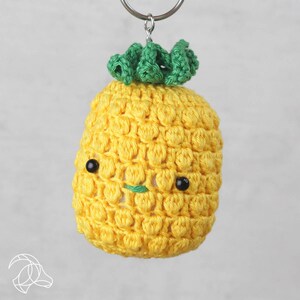 Könnte beinhalten: Ein gelber gehäkelter Ananas-Schlüsselanhänger mit grünen Blättern und schwarzen Augen.