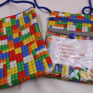 Könnte beinhalten: Zwei rechteckige Umhängetaschen mit einem bunten Muster aus ineinandergreifenden Kunststoff-Bausteinen in Rot, Gelb, Blau, Grün und Weiß. Jede Tasche hat ein blaues Kordelband. Eine Tasche enthält ein transparentes Fach mit einer weißen Karte.