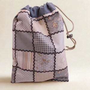 Könnte beinhalten: Ein Kordelzugbeutel mit Patchwork-Design. Die Tasche hat Quadrate mit verschiedenen Mustern, darunter Punkte, Hahnentritt, Blumen und Streifen, in Grau- und Beigetönen. Die Kanten der Quadrate sind mit dunkler Spitze verziert. Der obere Teil ist einfarbig blau-grau.