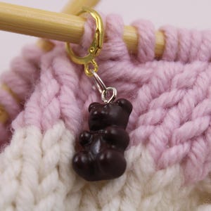 Könnte beinhalten: Ein brauner, bärenförmiger Maschenmarkierer mit einem goldenen Metallring, der an einem rosa und weißen gestrickten Stoff befestigt ist.