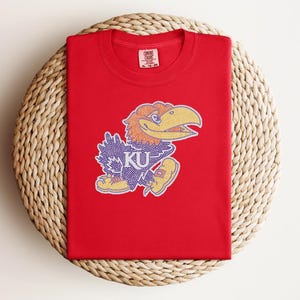 May include: A red t-shirt featuring a colorful cartoon Jayhawk mascot with the letters "KU". The Jayhawk has a yellow beak, orange hair, and purple and blue feathers. The shirt is folded on a woven, circular mat.