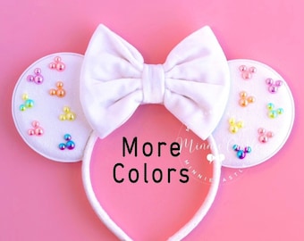 May include: White Minnie Mouse ears with a white bow and a white headband. The ears are decorated with colorful iridescent beads in the shape of Mickey Mouse heads. The text "More Colors" is printed on the headband.