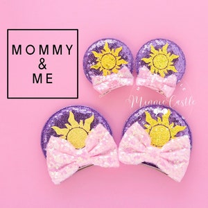 Könnte beinhalten: Zwei Paar violette und rosafarbene, mit Pailletten besetzte Minnie Mouse Ohren mit einem gelben Sonnendesign. Die Ohren sind mit einer rosafarbenen Schleife verziert und haben einen Clip auf der Rückseite. Der Text "Mommy & Me" ist auf dem Bild zu sehen.