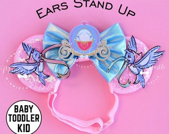 Peut inclure: Des oreilles de Minnie Mouse rose pailletées avec un nœud bleu et un charme de carrosse de Cendrillon. Les oreilles ont un oiseau bleu de chaque côté. Le bandeau est rose. Le texte "EARS STAND UP" est en haut de l'image. Le texte "BABY TODDLER KID" est en bas de l'image.