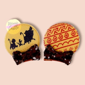 May include: A pair of brown and gold Minnie Mouse ears with a brown sequined bow. One ear features a silhouette of Simba, Nala, Timon, and Pumbaa from The Lion King. The other ear features a red and gold geometric pattern.