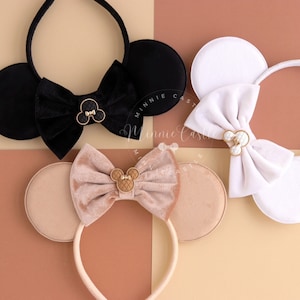 May include: Three pairs of mouse ears headbands with velvet bows and gold Mickey Mouse charms. The headbands are black, white, and beige.