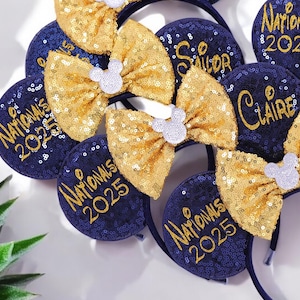 May include: A collection of navy blue sequined headbands with gold bows and white Mickey Mouse silhouettes. Some headbands have names like "Brooke", "Sailor", "Claire", and "Ella" in gold script. Other headbands have "Nationals 2025" in gold.