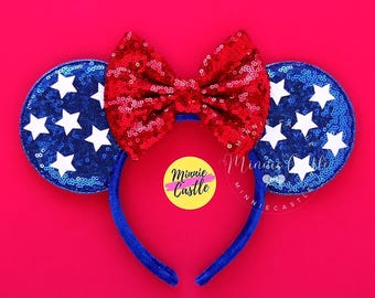Può includere: Orecchie di Minnie Mouse rosse, bianche e blu con un fiocco rosso di paillettes e orecchie blu con stelle bianche. Le orecchie sono su uno sfondo rosa.