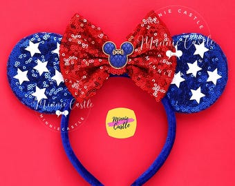 Può includere: Fascia per capelli con orecchie di Minnie Mouse rosso, bianco e blu con un fiocco rosso e stelle bianche sulle orecchie. La fascia è su uno sfondo rosso. Minnie Castle è scritto sulla fascia.