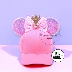 Puede incluir: Gorra de béisbol rosa con orejas de ratón con lentejuelas, un lazo rosa y una corona dorada. El lazo presenta un emblema de Minnie Mouse. La gorra tiene el texto "Minnie Castle" y una etiqueta "Kid Adult".