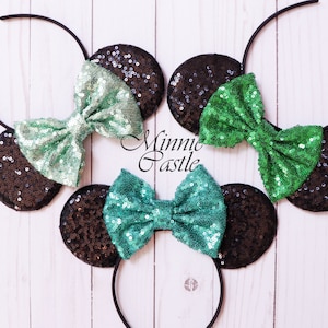 May include: Three black headband mouse ears with green and teal sequin bows. The headband is black and the ears are black with sequins. The bows are made of sequins and are tied in a bow shape. The text "Minnie Castle" is visible in the image.