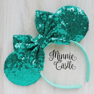 Puede incluir: Diadema de orejas de Minnie Mouse de lentejuelas color turquesa con un gran lazo. La diadema es de color verde menta y dice "Minnie Castle" en letras negras.