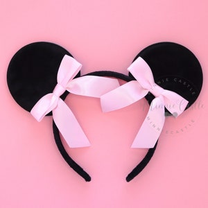 May include: Black velvet mouse ears headband with pink satin bows. The headband is on a pink background.