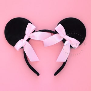 May include: Black mouse ear headband with two round ears and pink satin bows. The headband is on a pink background. The ears are made of a soft, velvety material. The bows are tied in the center of each ear.