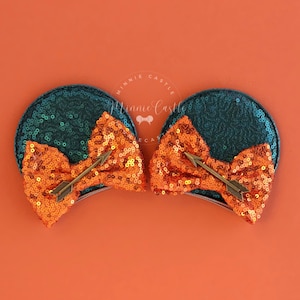 Op de afbeelding: Turquoise en oranje Minnie Mouse oren met een strik en pijl. De oren zijn bedekt met pailletten en de strik is gemaakt van glinsterend stof. De pijl is van goudkleurig metaal.