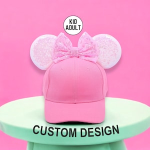 May include: Pink baseball cap with a sequined bow and ear accents. The cap has the text "CUSTOM DESIGN" at the bottom. The image also includes the text "KID ADULT" in a circle.