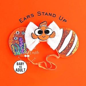 Peut inclure: Serre-tête avec oreilles à sequins orange, un nœud blanc pailleté et un personnage de poisson-clown. L'oreille gauche présente une broderie de corail bleu et violet avec des strass. Texte EARS STAND UP et BABY TO ADULT. Sangle orange incluse.