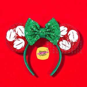 May include: A festive headband with red sequined ears featuring white floral designs. A large, sparkling green sequined bow sits atop a green headband. The background is a solid red.