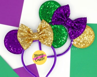May include: Purple and green sequined Minnie Mouse ears with a gold bow and headband. The headband is purple and the ears are decorated with gold, purple, and green sequins. The bow is gold and is tied in the centre of the headband. The text "Minnie Castle" is written on a yellow circle.