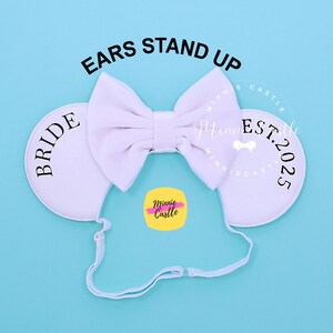 Può includere: Orecchie di Minnie Mouse bianche con un fiocco e la parola "BRIDE" su un orecchio e "Minnie Castle EST. 2025" sull'altro orecchio.