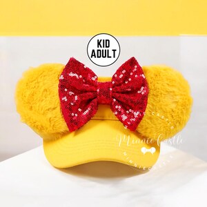May include: Yellow baseball cap with fuzzy yellow ears and a red sequined bow. The cap has the text "KID ADULT" on it and the text "Minnie Castle" on the brim.